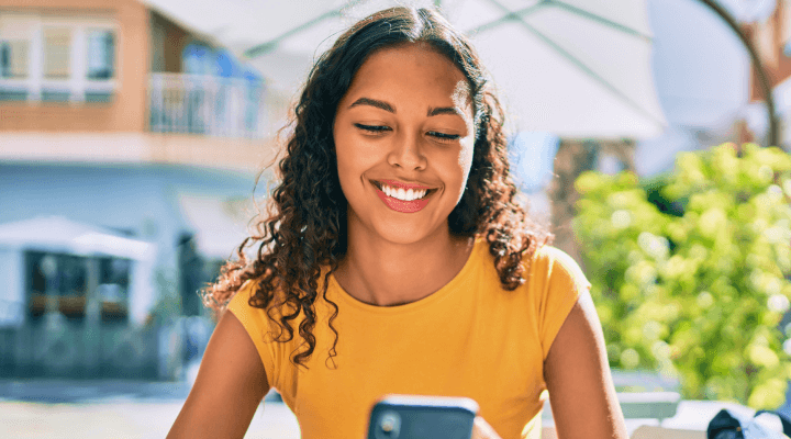 Num ambiente externo e ensolarado, uma jovem mulher sorri enquanto mexe no celular